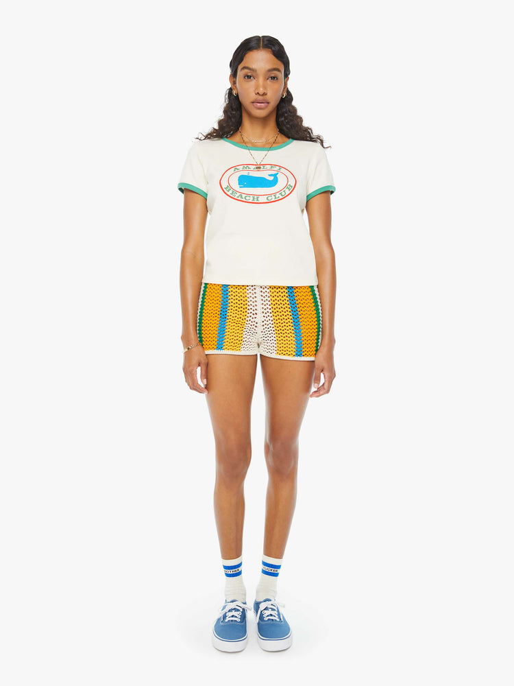 Full front view of a woman in an off-white shrunken crewneck tee with extra-short sleeves and a cropped hem designed with a blue whale graphic on the front and teal hems. Paired with crochet shorts and blue sneakers.