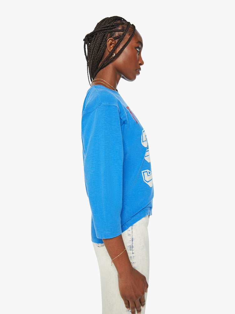 Side view of a women football tee with a boat neck, 3/4 sleeves, drop shoulders and a boxy fit in blue with number 32 and text in red.