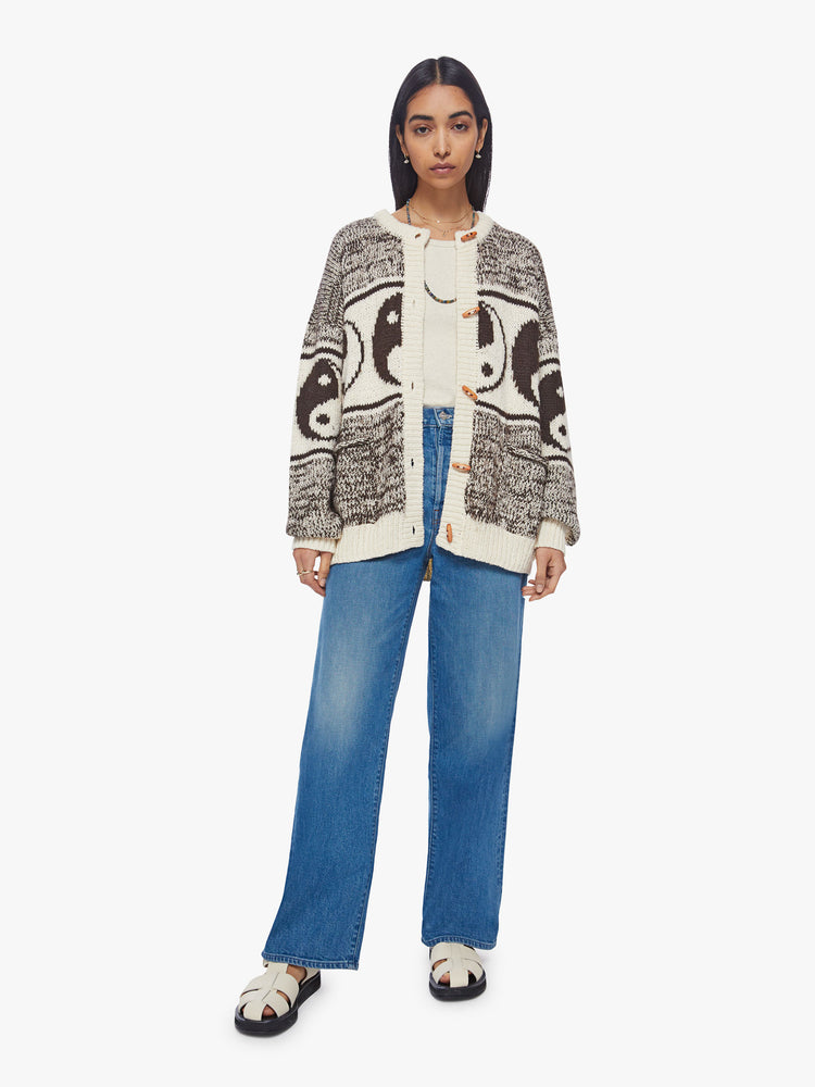 Full body view of a woman oversized crewneck cardigan with long balloon sleeves and ribbed hems in a marled brown and white knit with yin-yangs and horizontal stipes, wooden buttons.