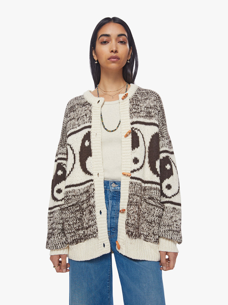 Front view of a woman oversized crewneck cardigan with long balloon sleeves and ribbed hems in a marled brown and white knit with yin-yangs and horizontal stipes, wooden buttons.
