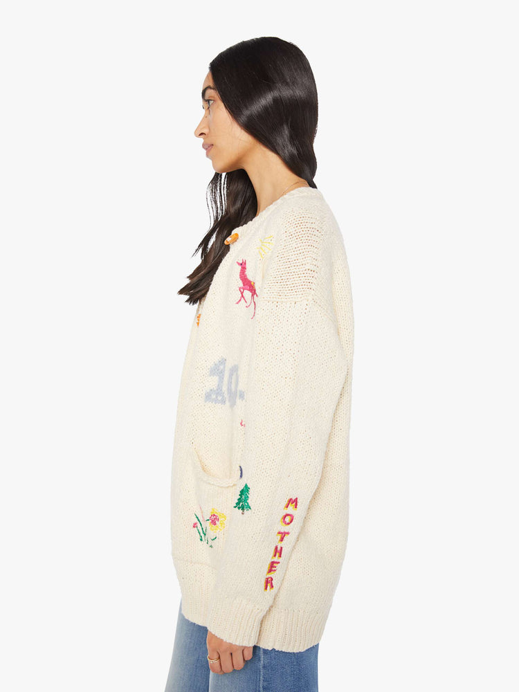 Side view of a woman in a cream with colorful embroidery  oversized crewneck cardigan with drop shoulders, patch pockets and ribbed hems. 