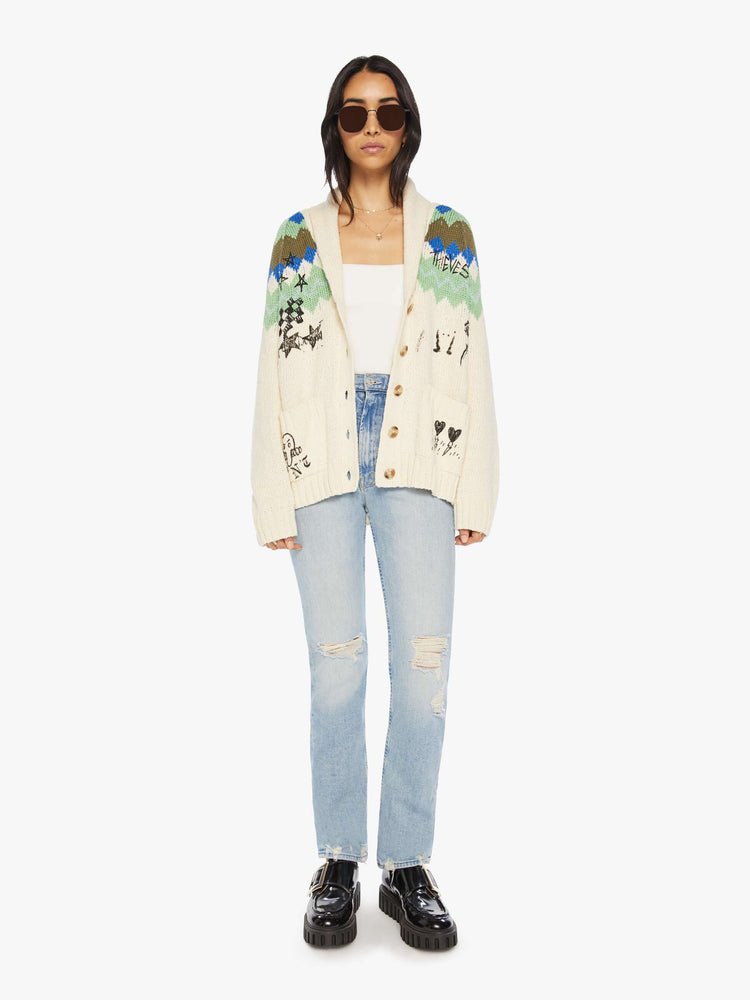 Full body view of a woman cardigan with a shawl collar, patch pockets, extra-long hems and buttons down the front in cream with a traditional knit pattern across the shoulders and angsty hand-drawn doodles inspired by bathroom graffiti.
