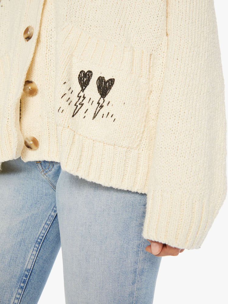 Close up swatch view of a woman cardigan with a shawl collar, patch pockets, extra-long hems and buttons down the front in cream with a traditional knit pattern across the shoulders and angsty hand-drawn doodles inspired by bathroom graffiti.