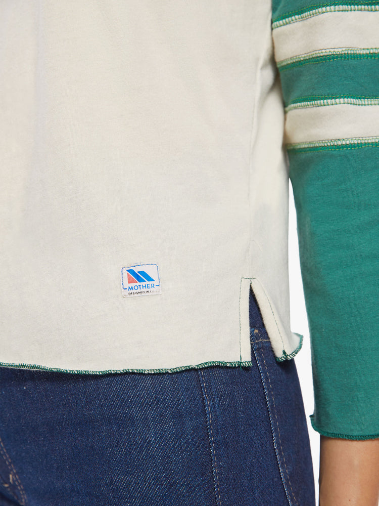 Close up hem view of a woman concert tee designed with a crew neck, 3/4 sleeves in an off-white hue with teal sleeves.