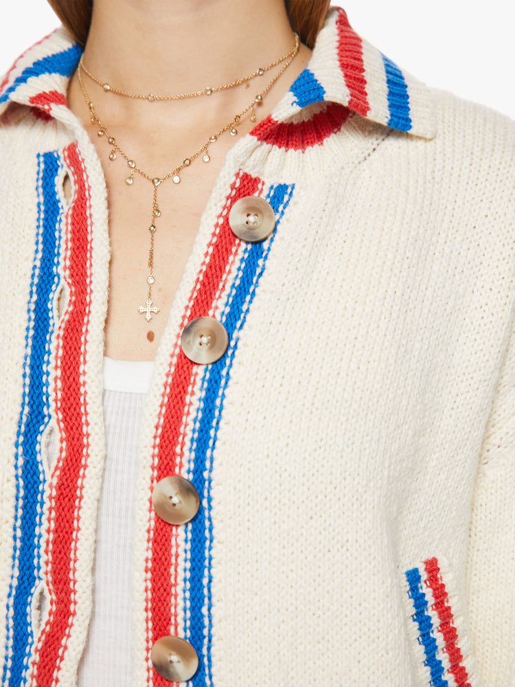 Close up view of a woman collared cardigan sweater with drop shoulders, slit pockets, thick ribbed hems and buttons down the front in cream features red and blue striped hems and athletic-inspired knit text and numbers on the back.
