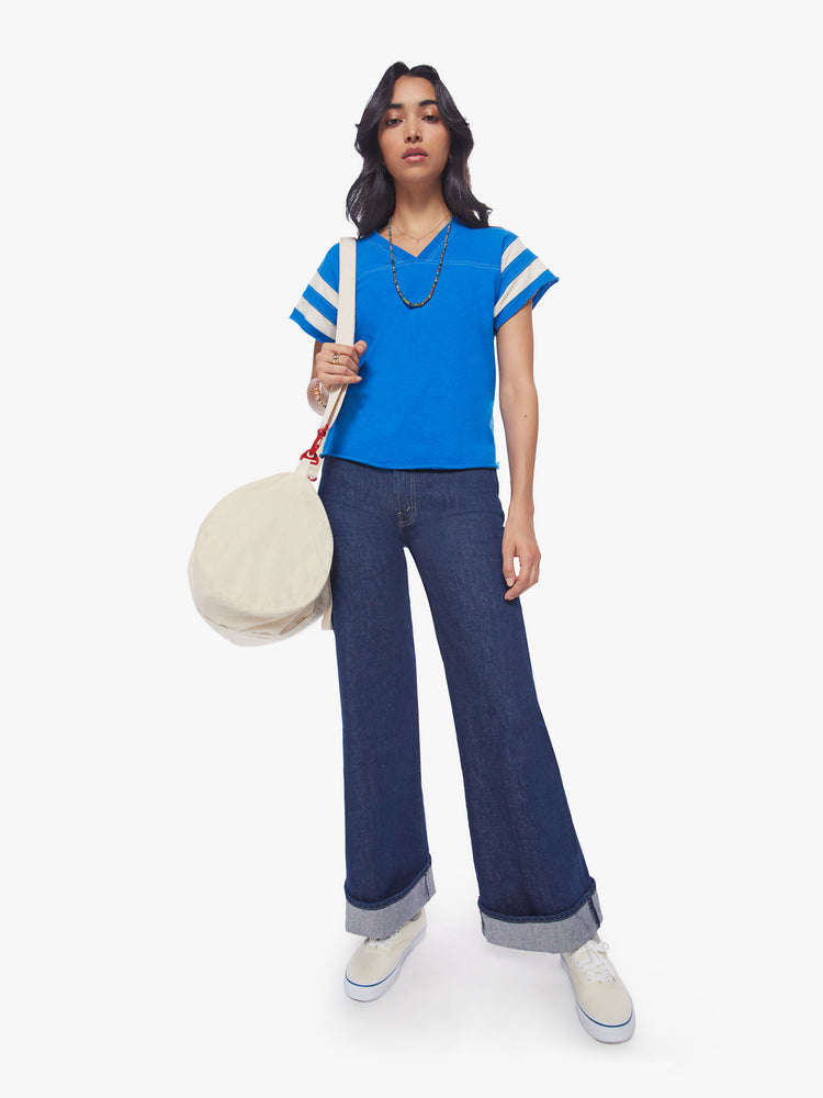 Full body view of a woman blue with white stripes on the sleeves football tee with a a V-neck with a seamed detail across the chest, short sleeves, drop shoulders.