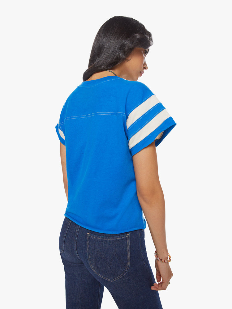 Back view of a woman blue with white stripes on the sleeves football tee with a a V-neck with a seamed detail across the chest, short sleeves, drop shoulders.