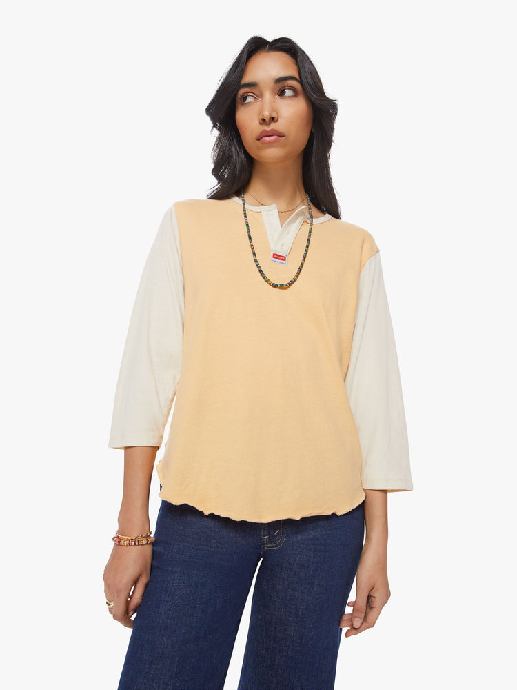 Front view of a woman in a 3/4 baseball style tee with a buttoned V-neck, curved hem and relaxed fit in a peach hue with off-white sleeves and a small red tag at the chest.