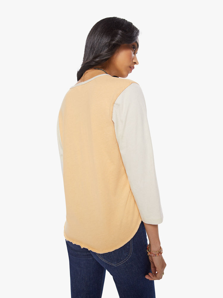 Back view of a woman in a 3/4 baseball style tee with a buttoned V-neck, curved hem and relaxed fit in a peach hue with off-white sleeves and a small red tag at the chest.