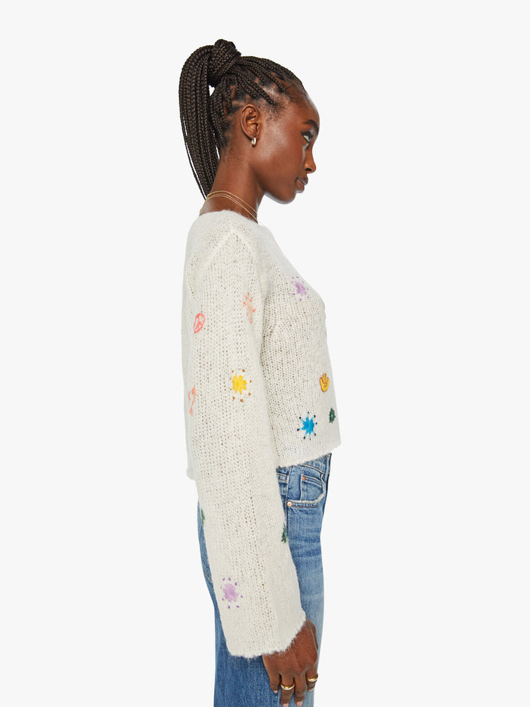 Side  view of woman boatneck sweater with long sleeves, drop shoulders, a cropped hem and boxy fit in an off white with colorful embroidered flowers, horses and other Western-inspired charms.