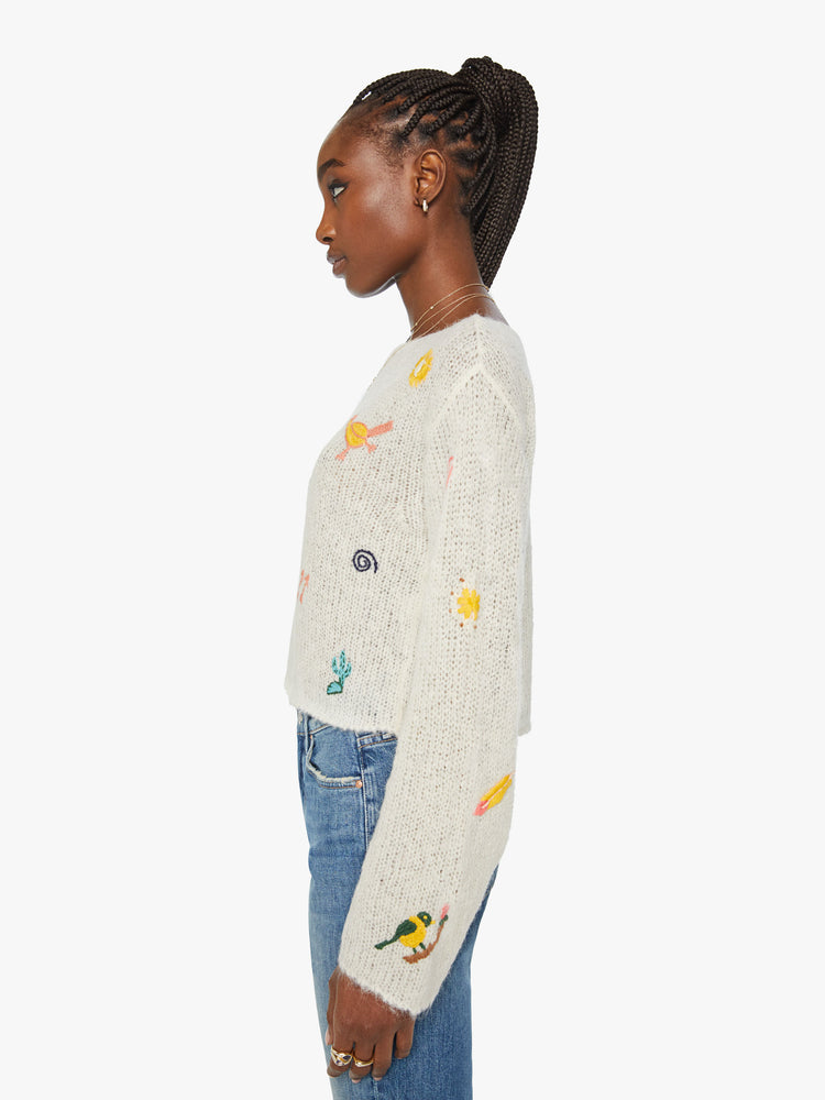 Left side view of woman boatneck sweater with long sleeves, drop shoulders, a cropped hem and boxy fit in an off white with colorful embroidered flowers, horses and other Western-inspired charms.