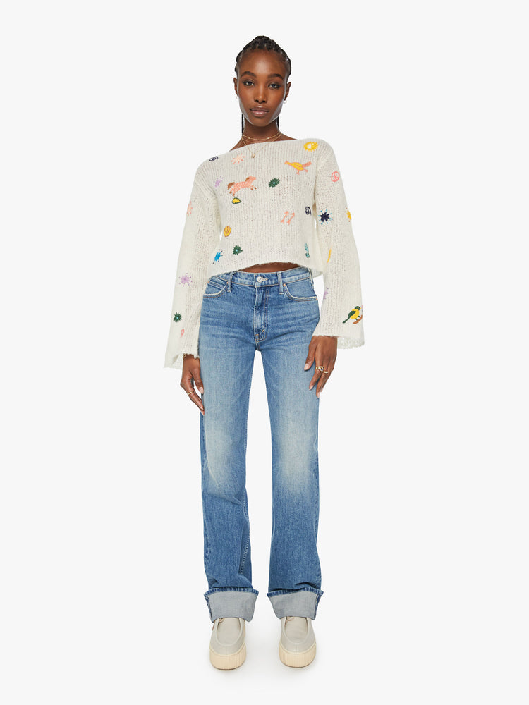 Full body  view of woman boatneck sweater with long sleeves, drop shoulders, a cropped hem and boxy fit in an off white with colorful embroidered flowers, horses and other Western-inspired charms.