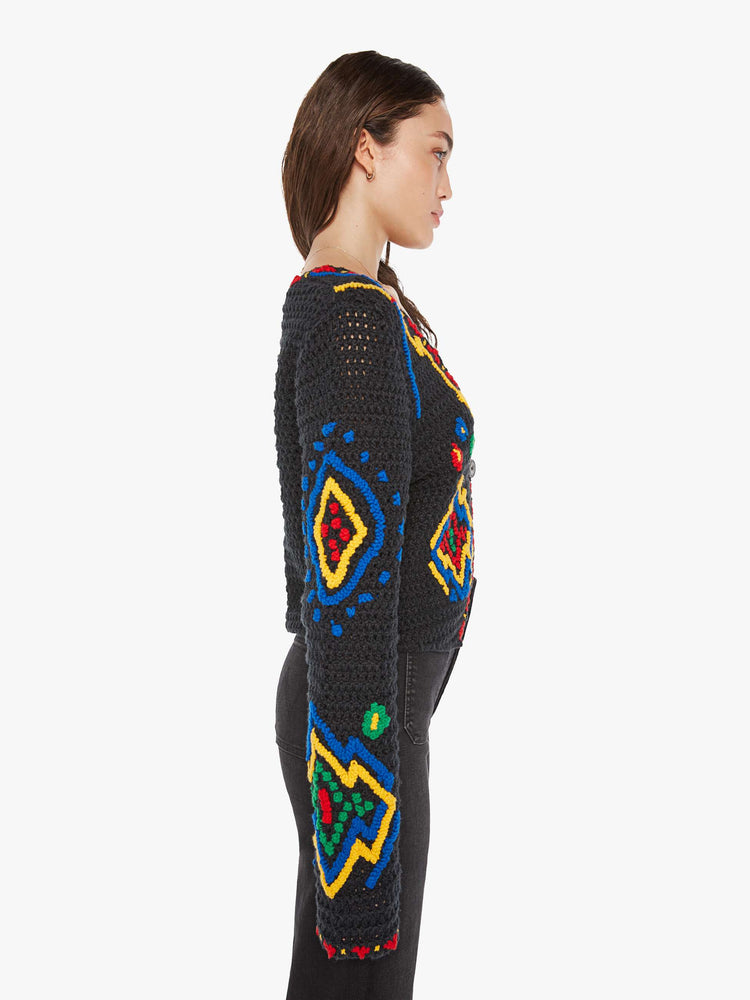 Side view of a woman in a black with colorful flowers, shapes and trim knit top with a deep V-neck, extra long sleeves and a single button closure.