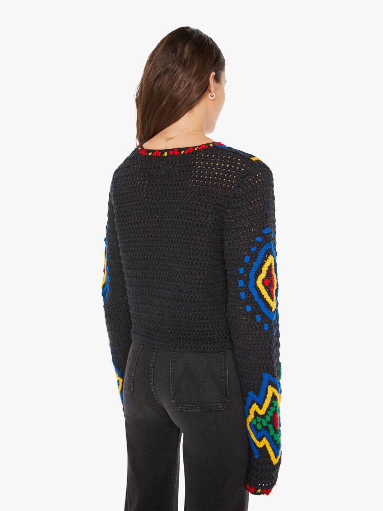 Back view of a woman in a black with colorful flowers, shapes and trim knit top with a deep V-neck, extra long sleeves and a single button closure.
