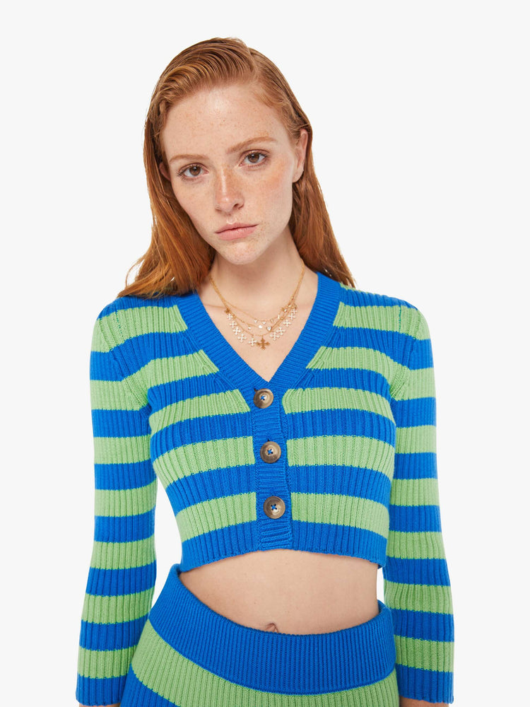 A front close up view of a woman wearing a green and blue striped knit cardigan featuring three brown buttons and a cropped body.