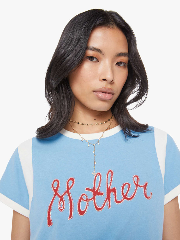 Close up view of a woman in a baby blue with white trim and red cursive  patch in the front tee with extra-short sleeves, inset details at the shoulders and a loose, boxy fit. 
