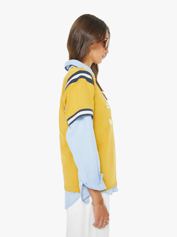 Side view of a woman in yellow tee features navy and white stripes at the neck, shoulder and sleeves, and a white text appliqued on the front inspired by a sports jersey.