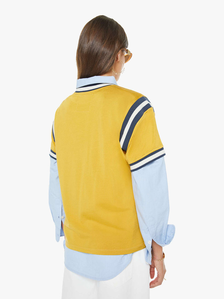 Back view of a woman in yellow tee features navy and white stripes at the neck, shoulder and sleeves, and a white text appliqued on the front inspired by a sports jersey.