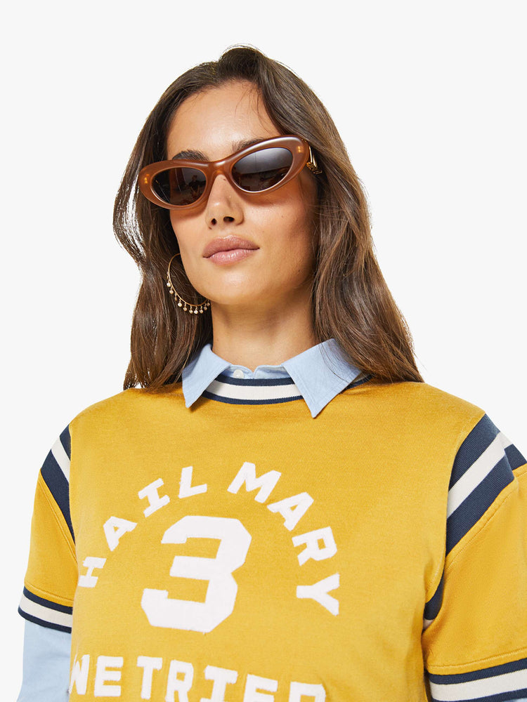 Close up view of a woman in yellow tee features navy and white stripes at the neck, shoulder and sleeves, and a white text appliqued on the front inspired by a sports jersey.