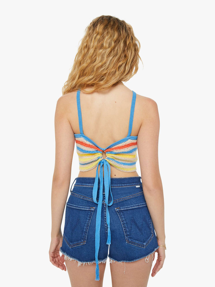 Back view of a woman in cream and rainbow striped crochet tank top that has narrow straps, a scoop neck and a cinch in the back that ties with openwork details and a flower motif at the chest.