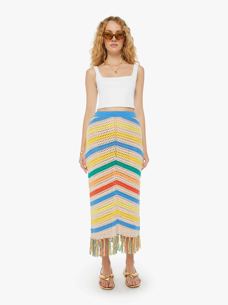 Front view of a woman in a high-waisted crochet cream and rainbow striped maxi skirt with a thick waistband and a fringed hem and a white tank top.