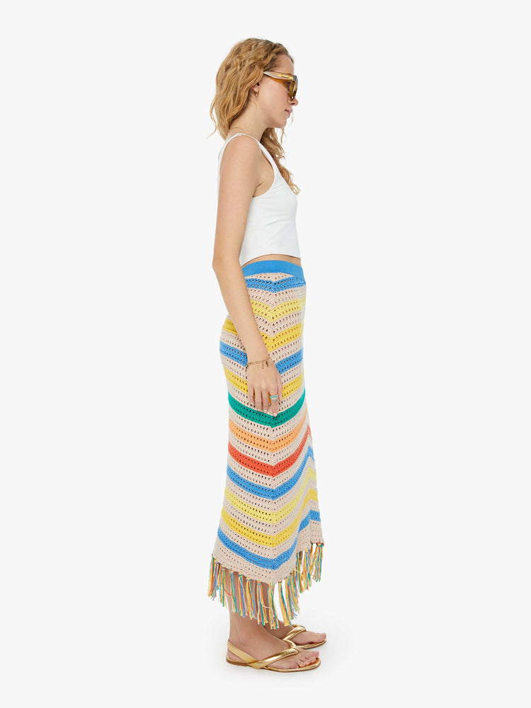 Side view of a woman in a high-waisted crochet cream and rainbow striped maxi skirt with a thick waistband and a fringed hem and a white tank top.