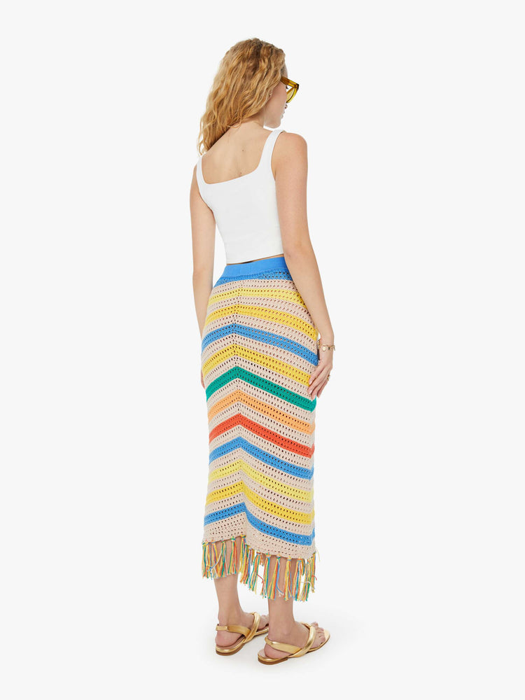 Back view of a woman in a high-waisted crochet cream and rainbow striped maxi skirt with a thick waistband and a fringed hem and a white tank top.