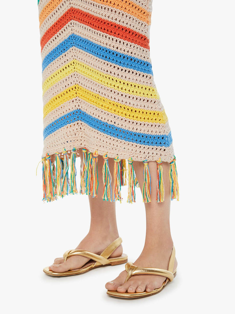 Swatch view of a woman in a high-waisted crochet cream and rainbow striped maxi skirt with a thick waistband and a fringed hem with gold sandals