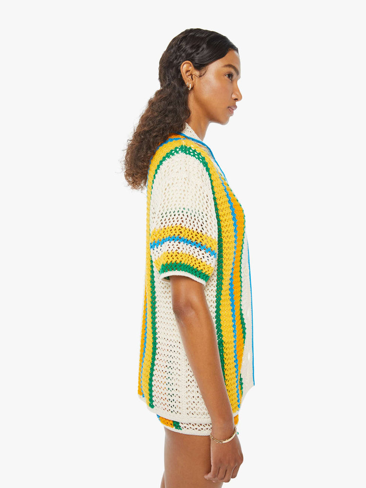 Side view of a woman in a cream, orange, green and blue striped crochet shir twith drop shoulders and a loose, oversized fit featuring delicate openwork details.