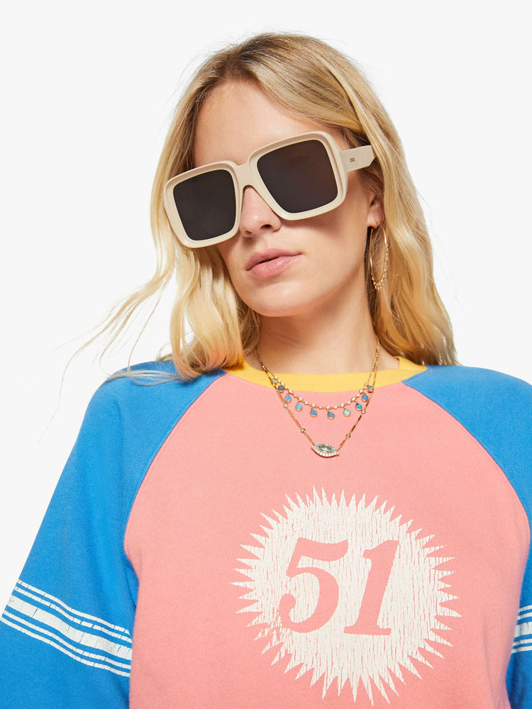 Close up view of a woman in a pale pink with blue sleeves and yellow trim sweatshirt and features a crackle-printed 51 on the chest and white stripes on the sleeves.