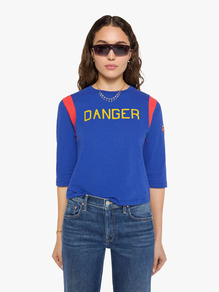 Front view of a woman in a primary blue tee with red inserts at the shoulders, a flocked text graphic in yellow across the chest and an embroidered tiger on the sleeve.