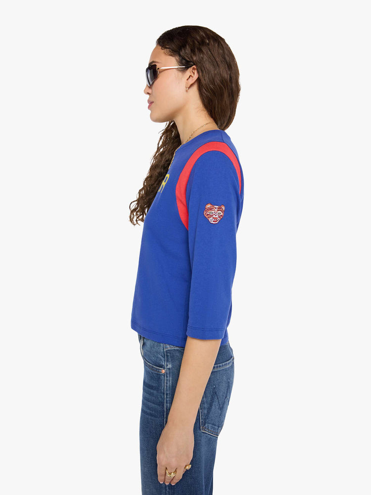 Side view of a woman in a primary blue tee with red inserts at the shoulders, a flocked text graphic in yellow across the chest and an embroidered tiger on the sleeve.