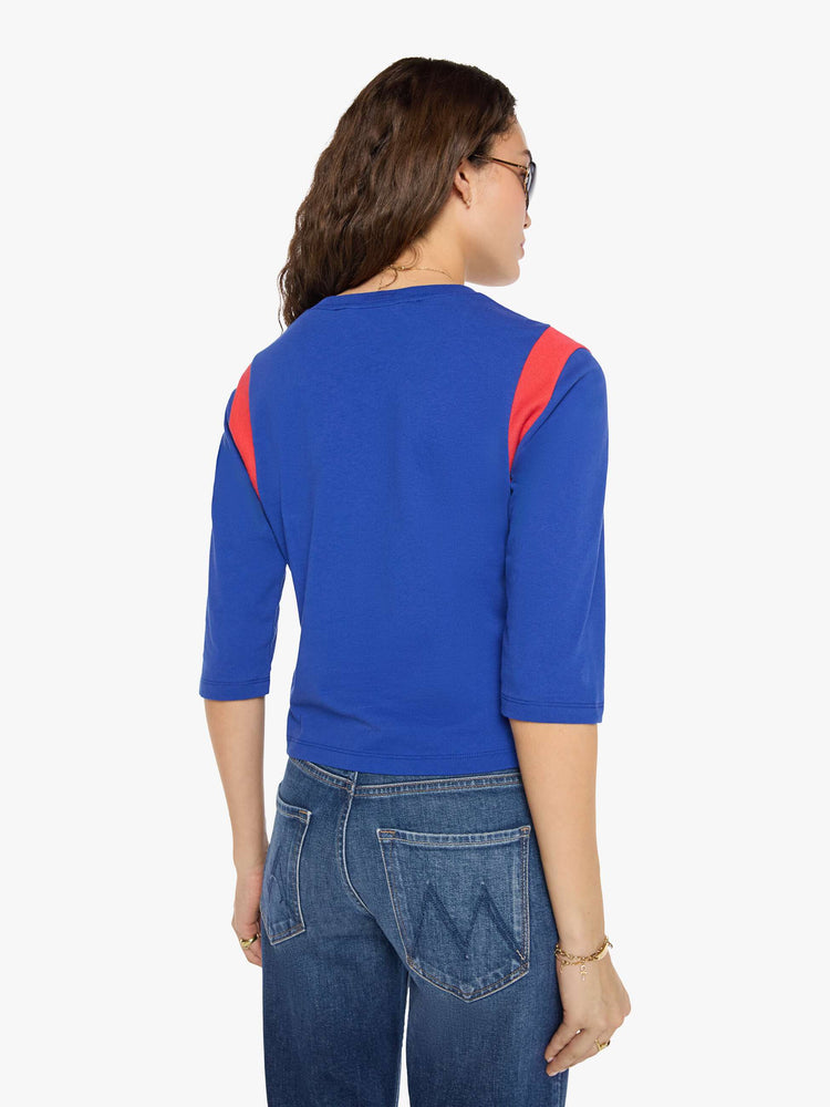 Back view of a woman in a primary blue tee with red inserts at the shoulders, a flocked text graphic in yellow across the chest and an embroidered tiger on the sleeve.
