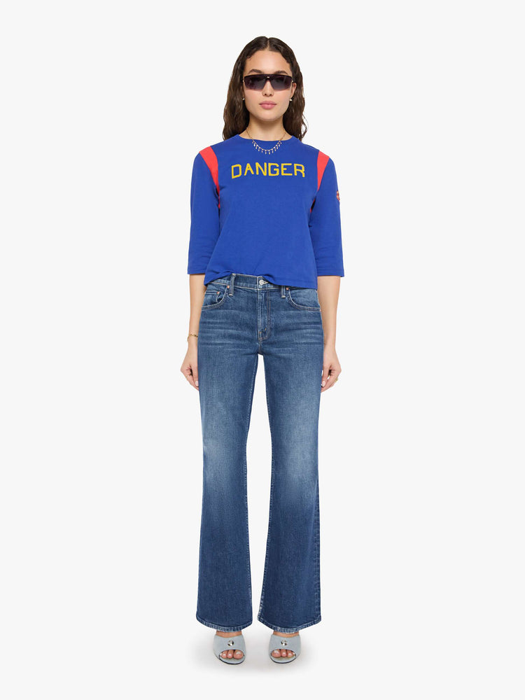 Full body view of a woman in a primary blue tee with red inserts at the shoulders, a flocked text graphic in yellow across the chest and an embroidered tiger on the sleeve.
