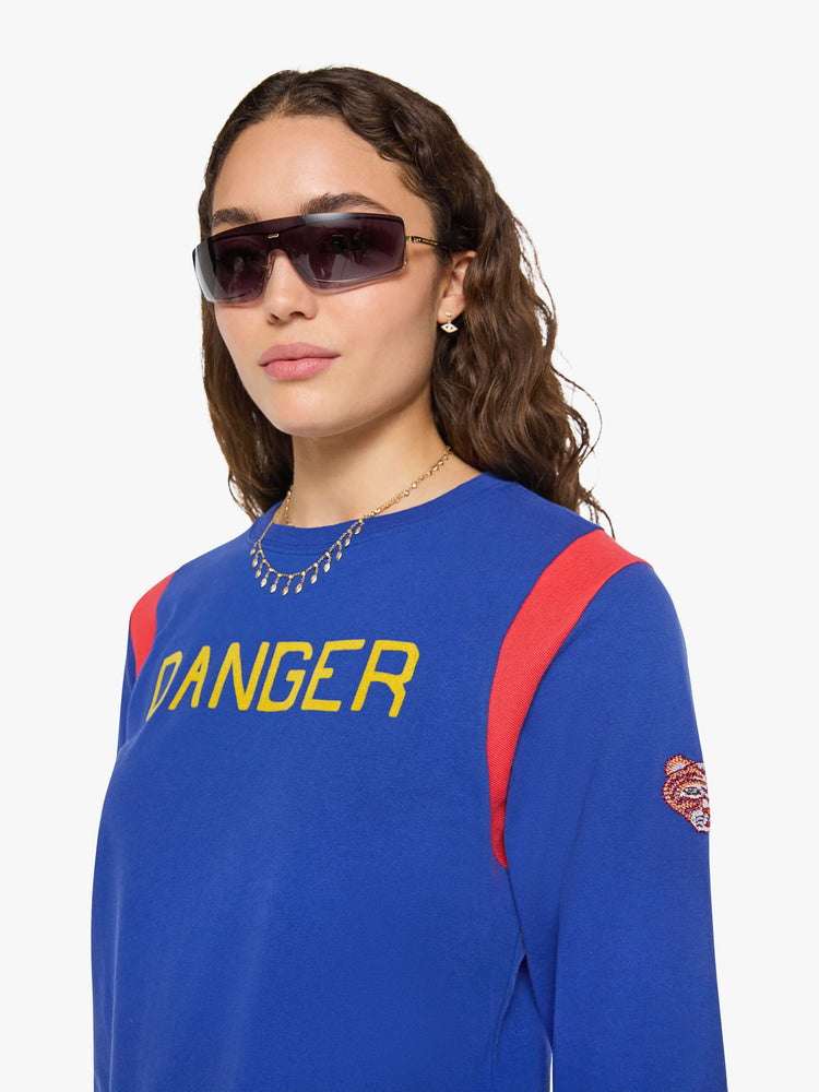 Close up view of a woman in a primary blue tee with red inserts at the shoulders, a flocked text graphic in yellow across the chest and an embroidered tiger on the sleeve.