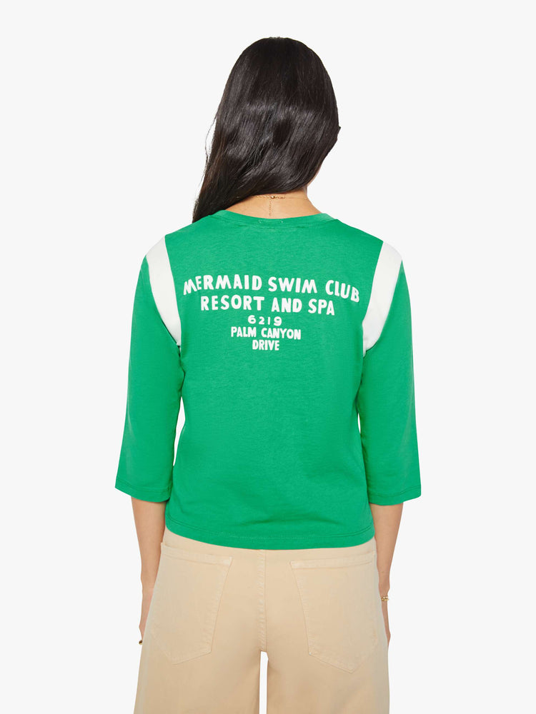 Back view of a woman in green with white inserts at the sleeves tee with a flocked swim club graphic on the front and text in white on the back.