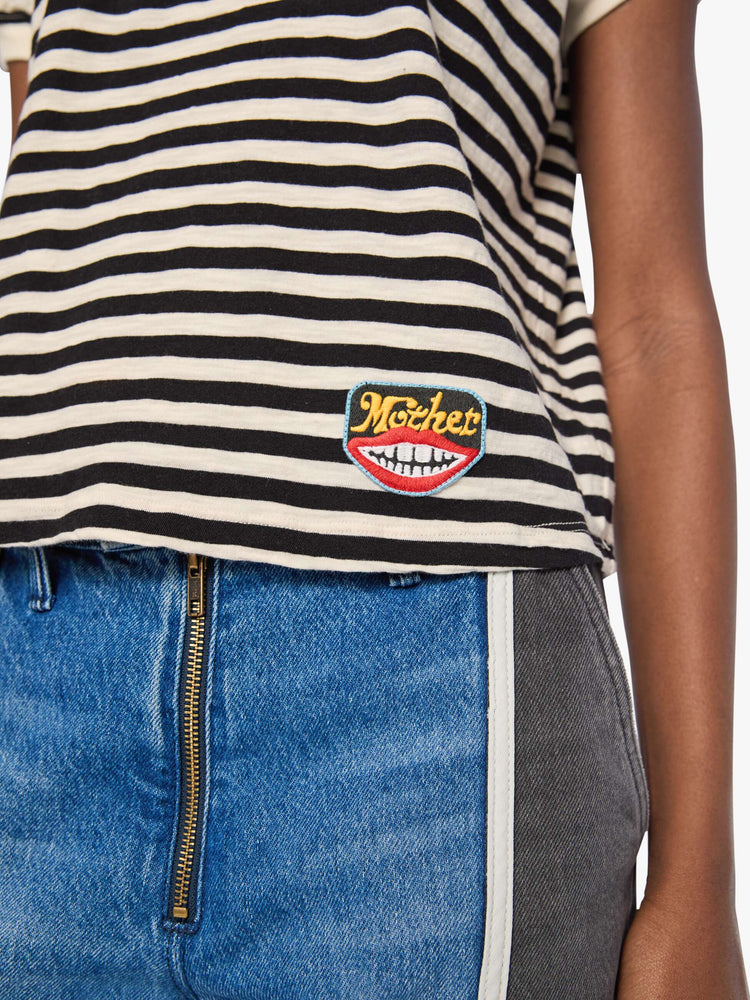 Close up view of a woman in a black and cream stripes tee features a mouth patch with MOTHER's name in yellow on the hip.