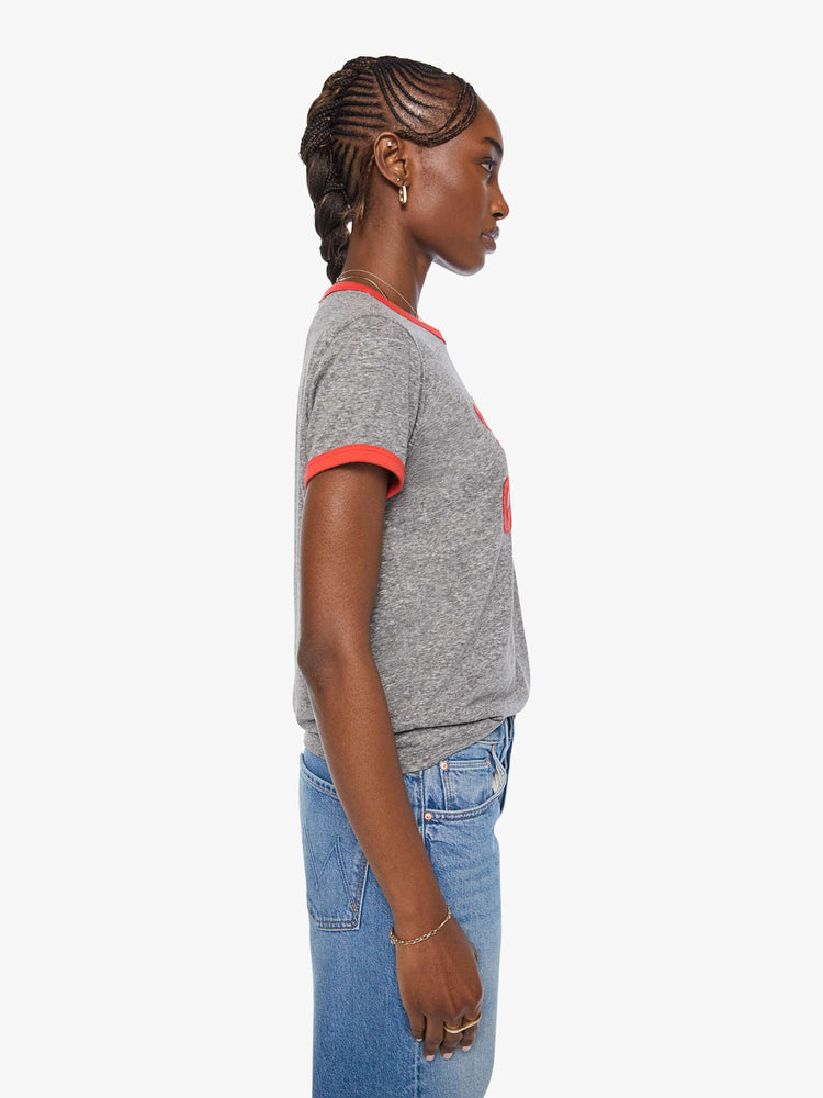 Side view of a woman in a heathered grey tee features red trim and an applique text graphic with small metal studs on the front.