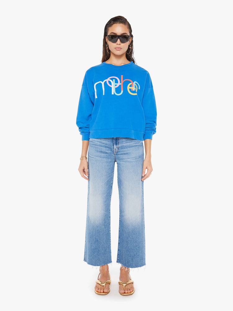 Front full body view of a woman wearing a bright blue oversized sweatshirt featuring a colorful graphic reading "mother", paired with a medium blue wash jean.