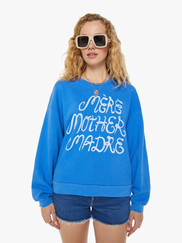 A front view of a woman wearing a blue sweatshirt featuring an oversized fit and a white graphic reading "MERE MOTHER MADRE", paired with dark blue denim short.