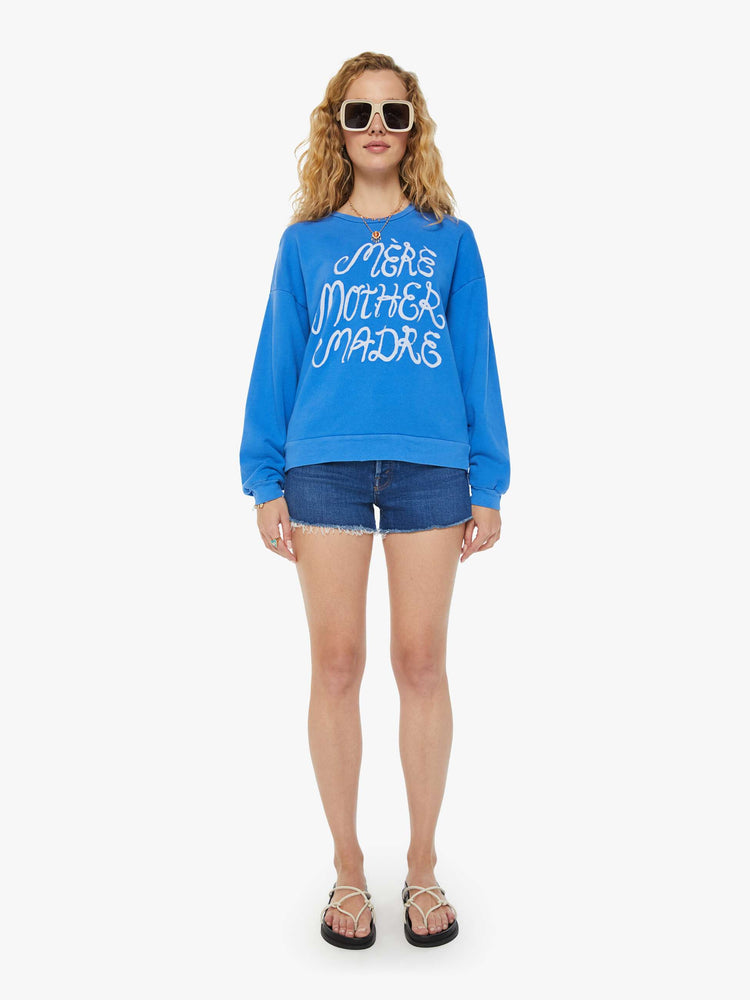 A front full body view of a woman wearing a blue sweatshirt featuring an oversized fit and a white graphic reading "MERE MOTHER MADRE", paired with dark blue denim short.