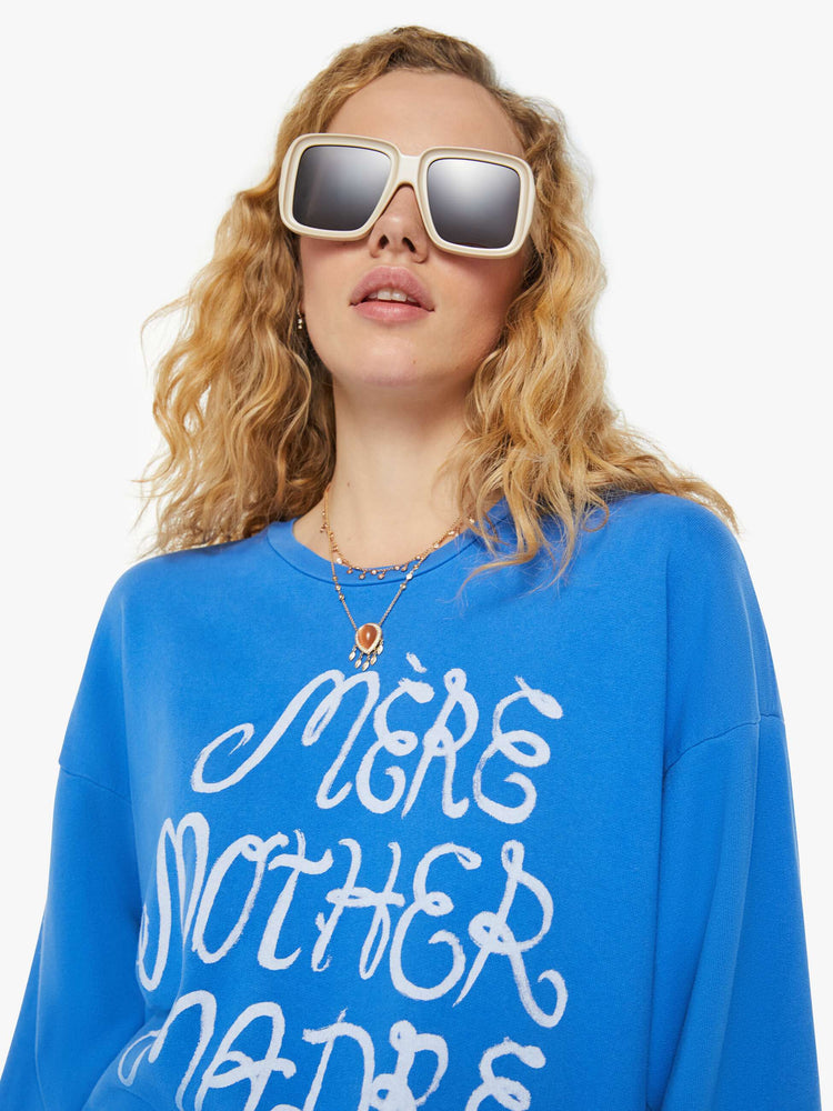 A front close up view of a woman wearing a blue sweatshirt featuring an oversized fit and a white graphic reading "MERE MOTHER MADRE".