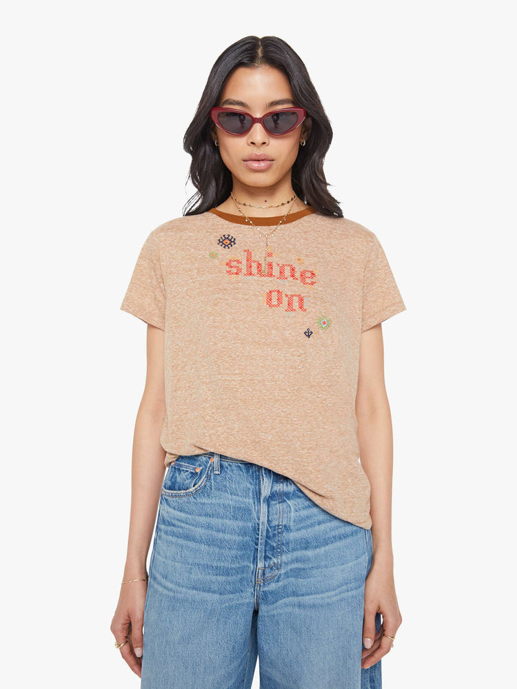 Front view of a woman in a heathered tan tee with dark brown trim along the neck, the tee features red text and colorful folk-inspired embroidery throughout.