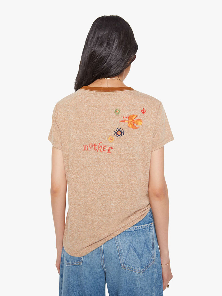 Back view of a woman in a heathered tan tee with dark brown trim along the neck, the tee features red text and colorful folk-inspired embroidery throughout.