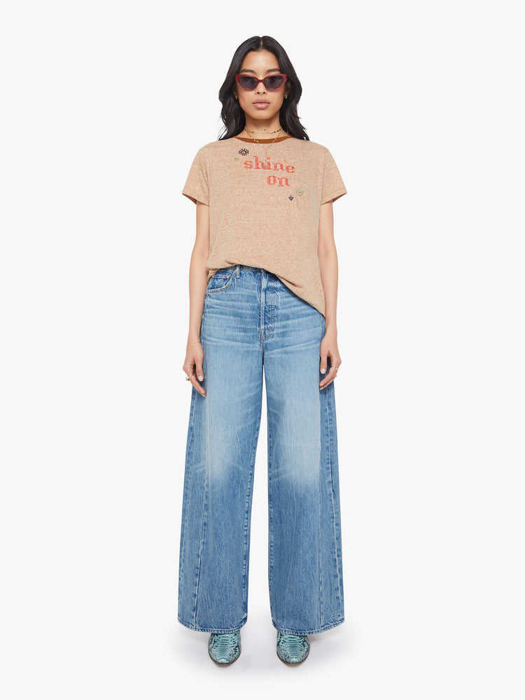 Full body view of a woman in a heathered tan tee with dark brown trim along the neck, the tee features red text and colorful folk-inspired embroidery throughout.