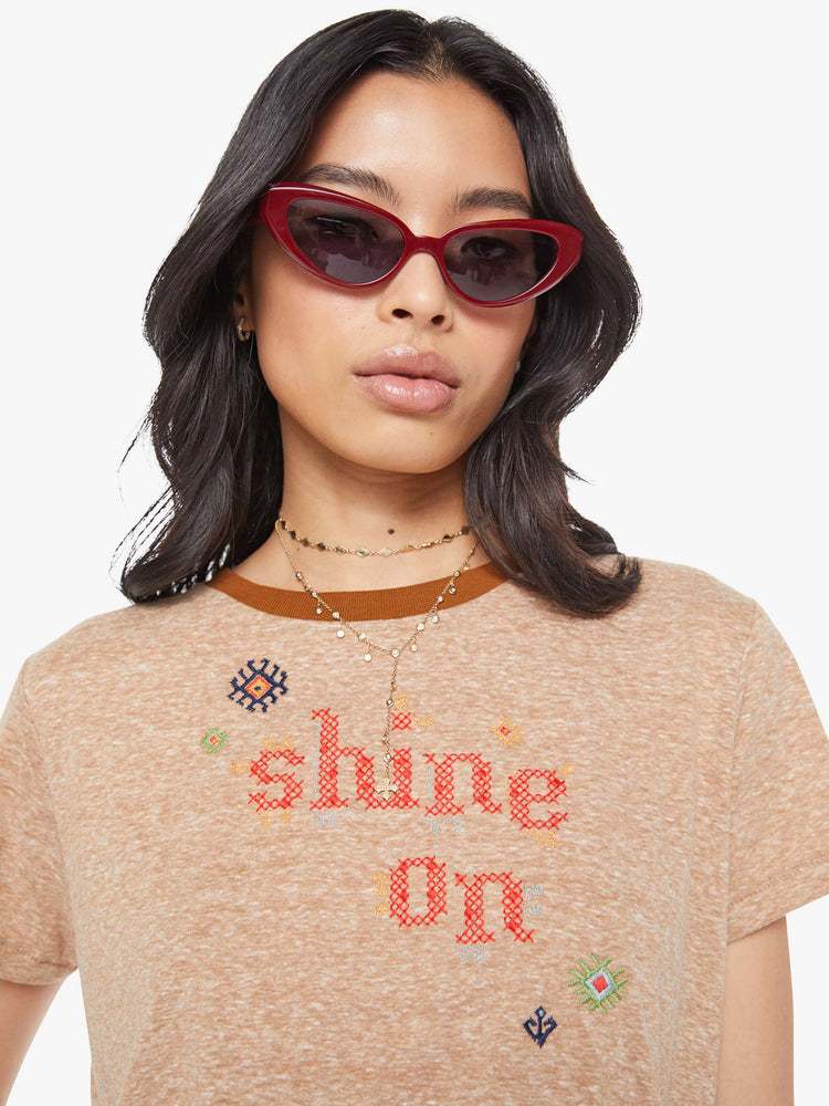 Close up view of a woman in a heathered tan tee with dark brown trim along the neck, the tee features red text and colorful folk-inspired embroidery throughout.