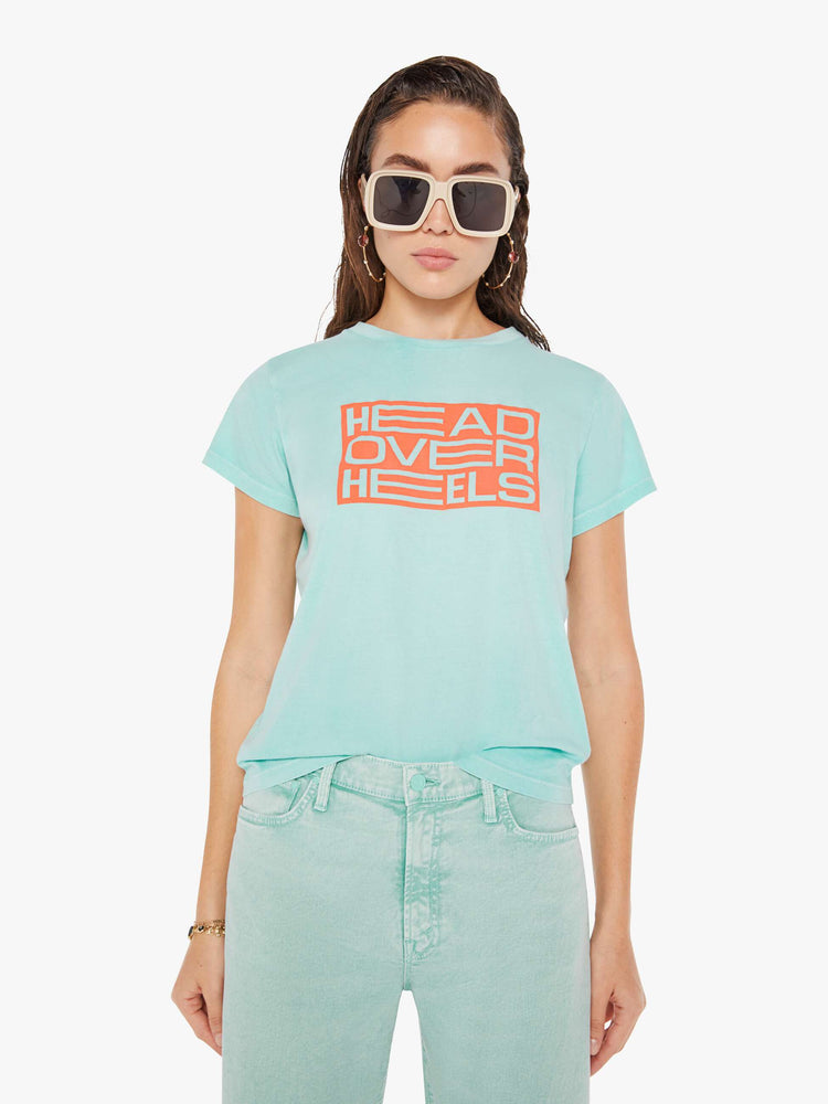 Front view of a woman wearing a light teal, fitted crew neck tee, featuring an orange rectangular graphic reading "HEAD OVER HEELS", paired with a light teal pant.