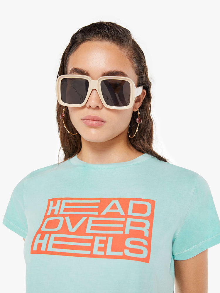 Front close up view of a woman wearing a light teal, fitted crew neck tee, featuring an orange rectangular graphic reading "HEAD OVER HEELS".