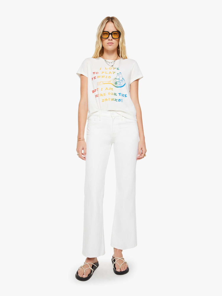 Full body view of a woman in a white tee that features  a multi-color text graphic with a tennis racket and cocktails on the front.