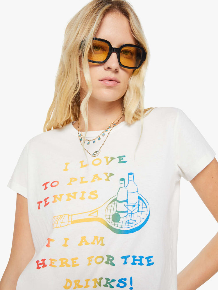 Close up view of a woman in a white tee that features  a multi-color text graphic with a tennis racket and cocktails on the front.